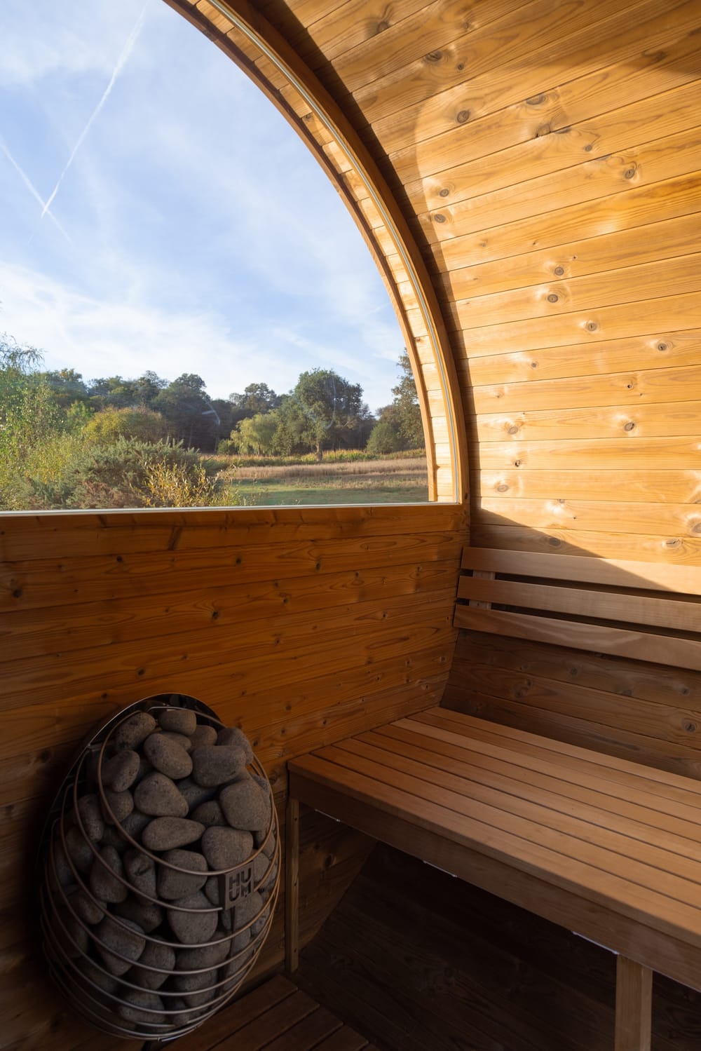 Inside the sauna: timber-lined walls, a bench, a heater piled with stones and a half-round window onto sky and trees