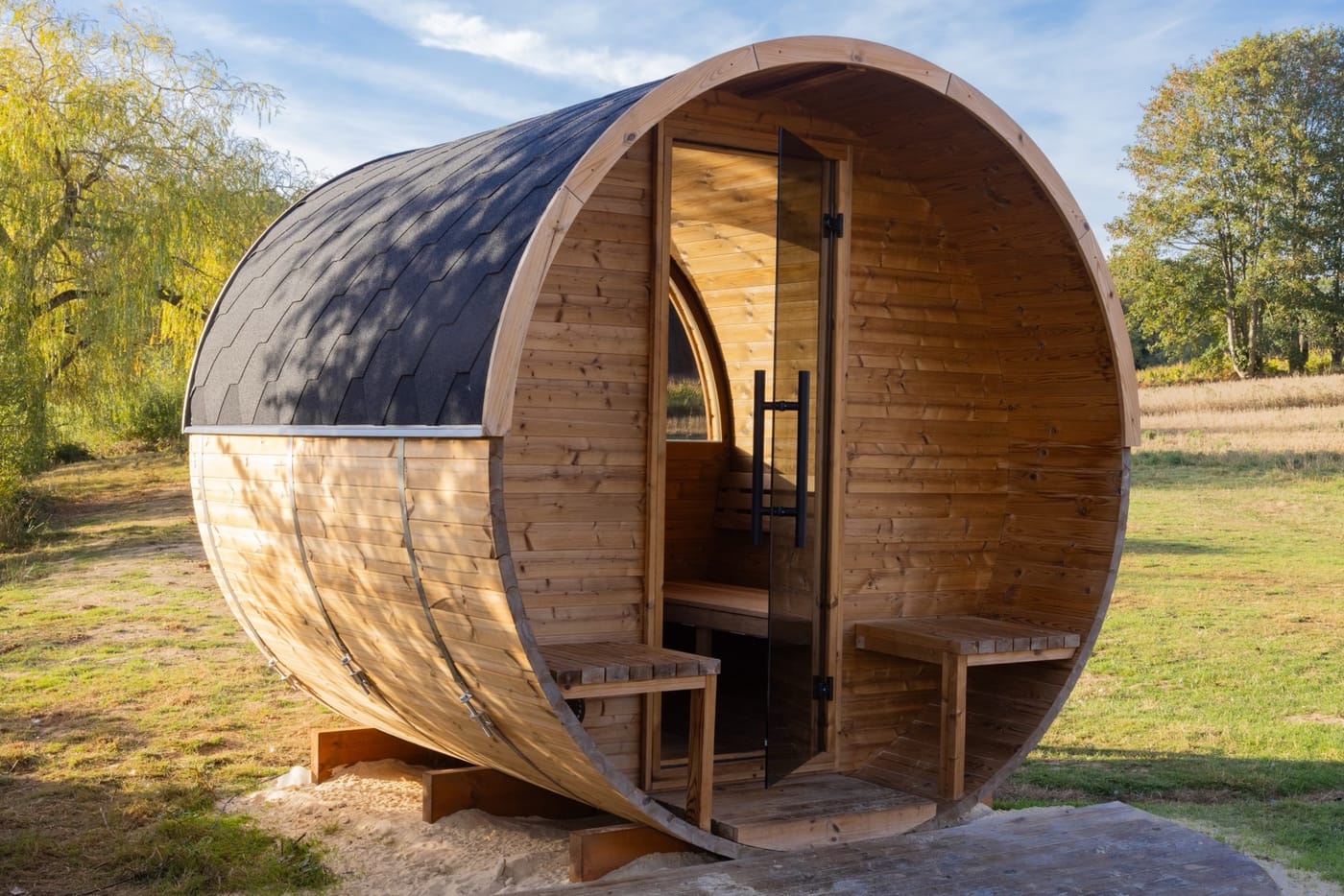 A barrel sauna on grass in morning light: a round timber front, a dark shingled roof, a glass door standing open and a small bench either side of it